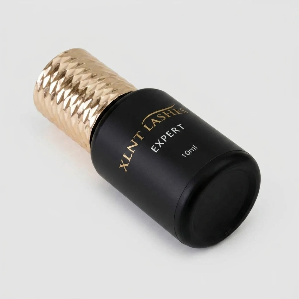 Svart XLNT Lashes Expert Glue 10ml 1 Sek flaska med mönstrat guldlock liggande på ljus bakgrund.