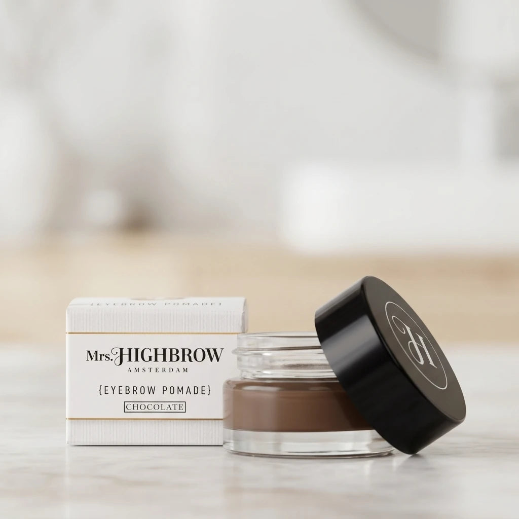 Mrs. Highbrow Eyebrow Pomade Chocolate i glasburk bredvid vit förpackning på ljus marmoryta, sidovy.