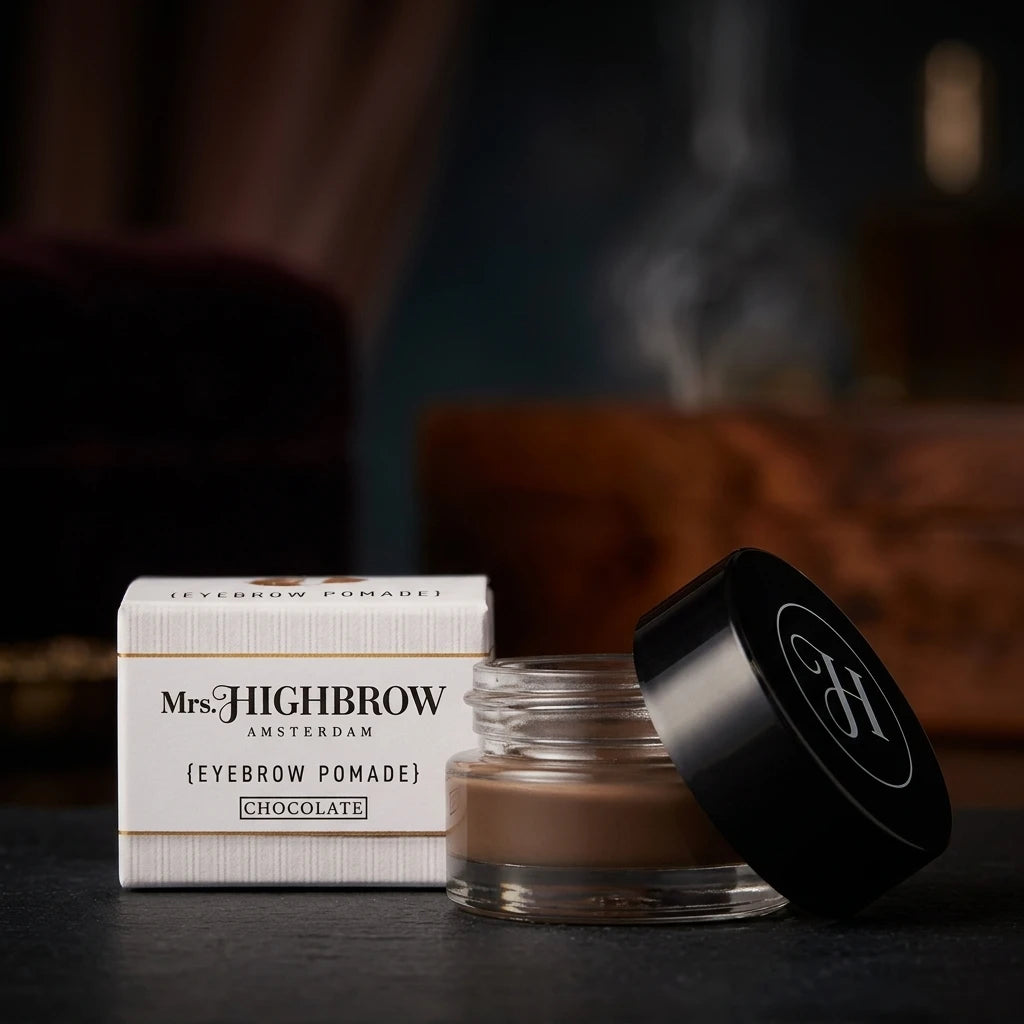Mrs. Highbrow Eyebrow Pomade Chocolate i glasburk med lutat svart lock bredvid vit förpackning på mörkt underlag.