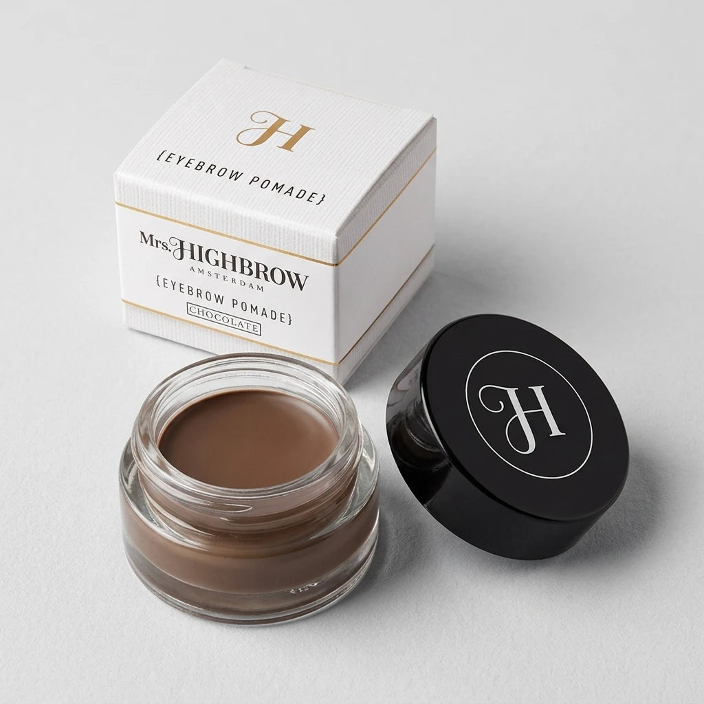 Mrs. Highbrow Eyebrow Pomade i liten glasburk med öppet svart lock framför vit pappersförpackning på grå bakgrund.