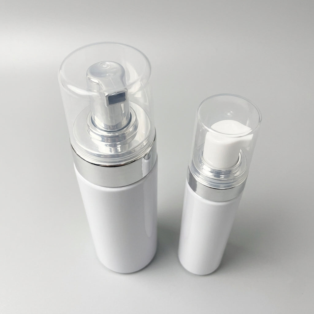 Vit cylinderformad Foam Flaska 75 ml med silverpump och genomskinligt lock bredvid en mindre flaska på grå bakgrund.