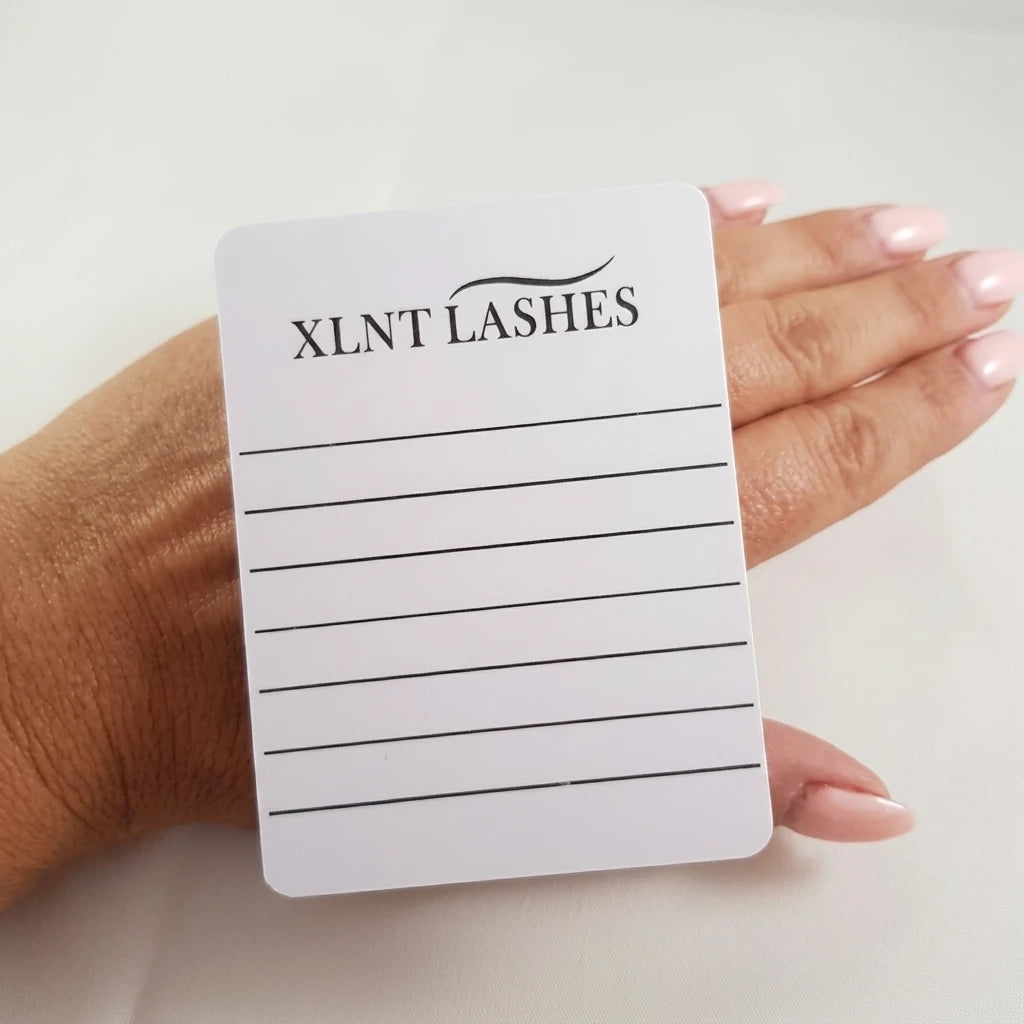 Vit akrylfransplatta från XLNT Lashes fäst på handrygg med resårband, rosa naglar synliga.