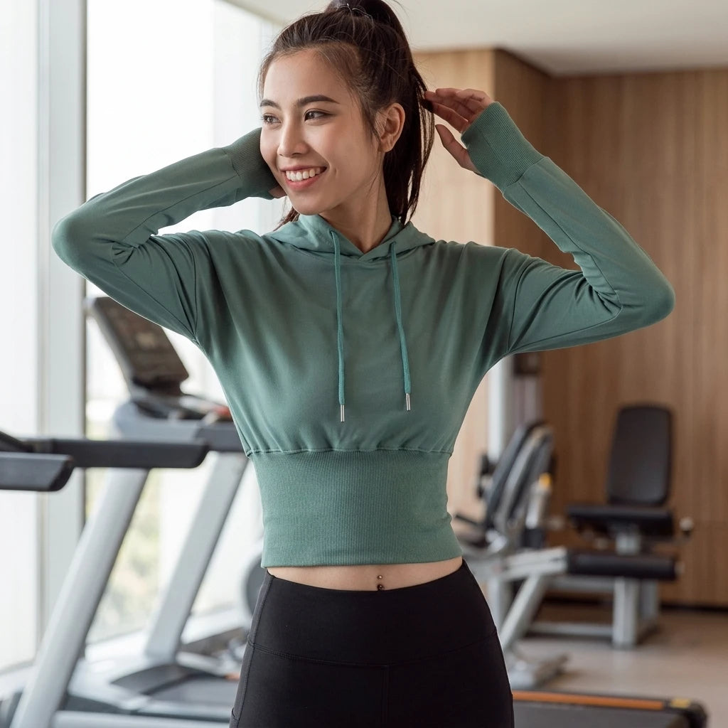 Leende kvinna på gym bär grön croppad hoodie med ribbad midja och dragsko, stylade med svarta träningstights.