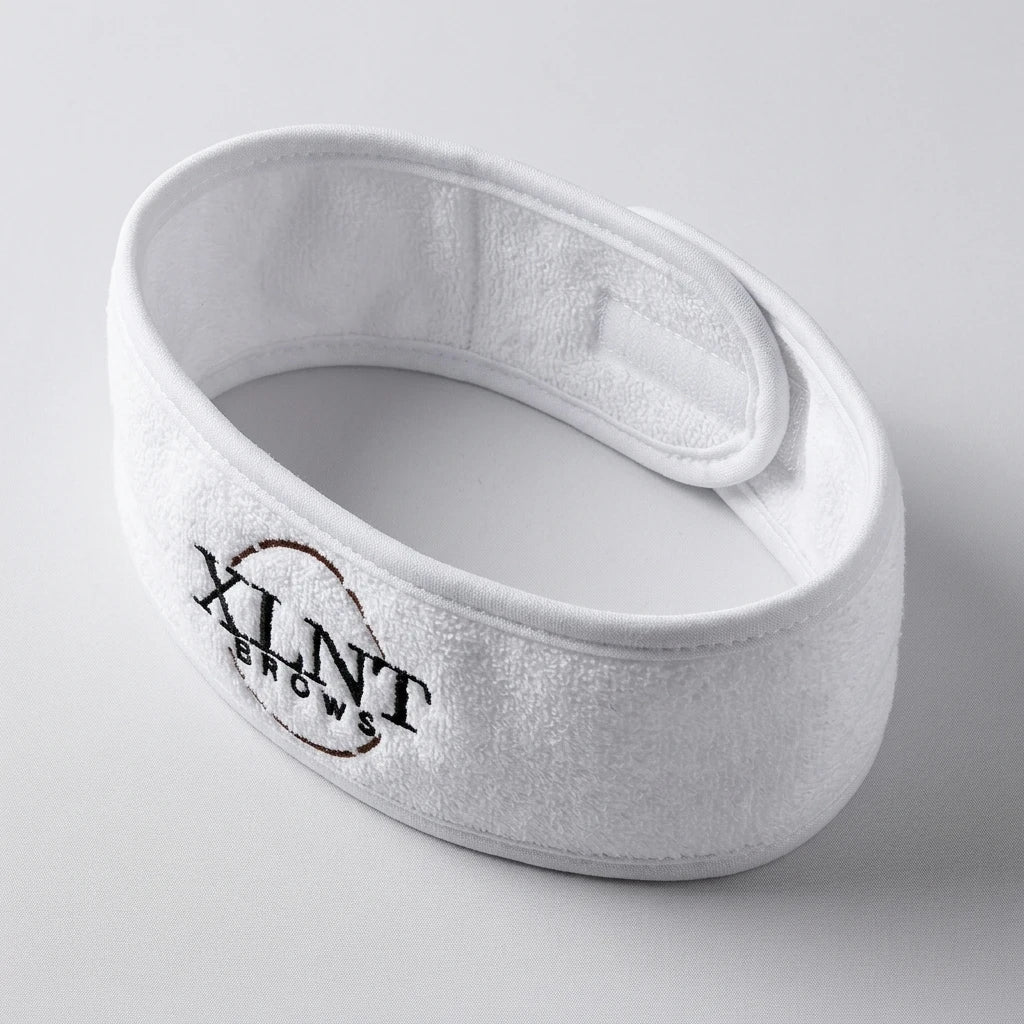 Brett vitt frottéhårband med kardborreknäppning och broderad XLNT Brows-logotyp, ovanifrån.