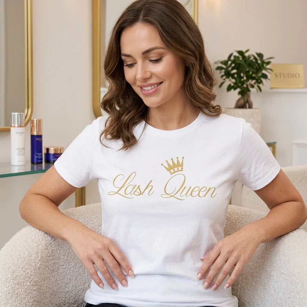 Kvinna i vit Lash Queen T-shirt med guldtryck och krona, sittande avslappnat i en ljus salongsmiljö.