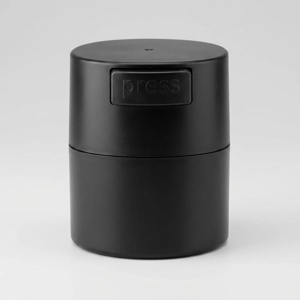 Svart cylinderformad Glue Box i matt plast med pressknapp på locket, mot ljusgrå bakgrund.